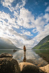 Seen from behind, a person stands in a lake on a sunny day. Two sloped mountains are in the distance in front of the person.