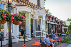 People walk into a café on a summer day from a street in a historic downtown. The building has wide windows and white façade.