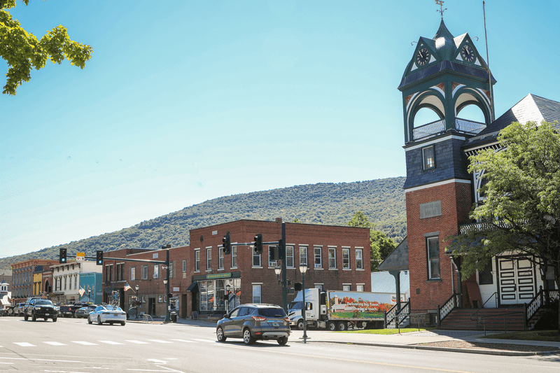 Brick buildings line a street with cars and mountains in the background on a sunny summer day.