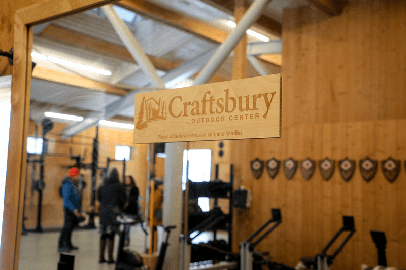 Indoor scene of a ski lodge and a sign that reads Craftsbury and people in the background getting fitted for skis.