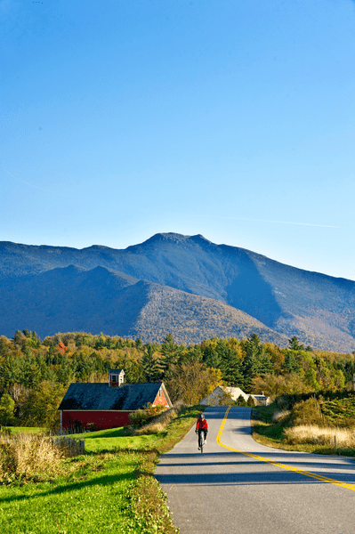 A person bikes on a paved road on a sunny day. A barn and mountains sit in the background.