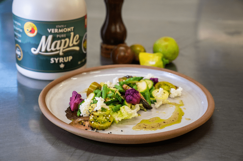 Colorful vegetables lined up on a plate with a jug of maple syrup behind the plate.