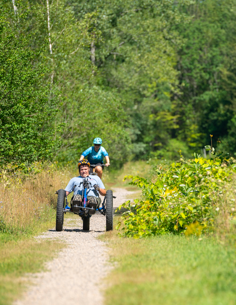 A person bikes using a three-wheeled adaptive bike on a gravel path.