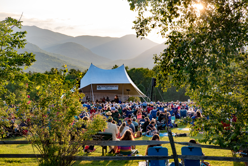 A crowd sits in front of an outdoor stage on a warm day.