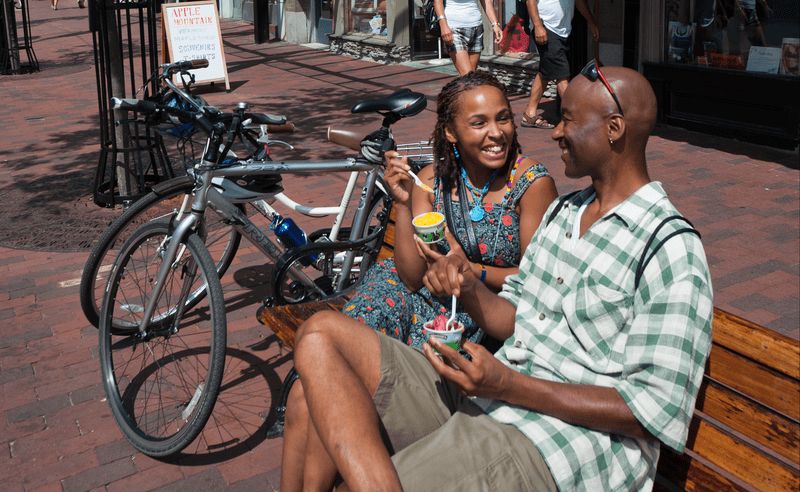 Two people enjoy ice cream from cubs outside on a park bench on a warm sunny day.