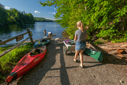 Several kayaks and canoes lay beached at a lake and are being loaded into the water.