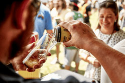 A person pours beer from a can into a glass in a tent surrounded by other people also holding beers and smiling.