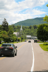 Cars drive along a road in a historic downtown with a white church in the background.