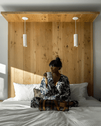 A person sits on a bed in a hotel room with a wool blanket rolled up in front of them.