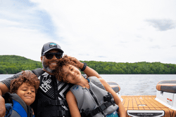 Three people are on a boat wearing personal flotation devices on a warm sunny day.