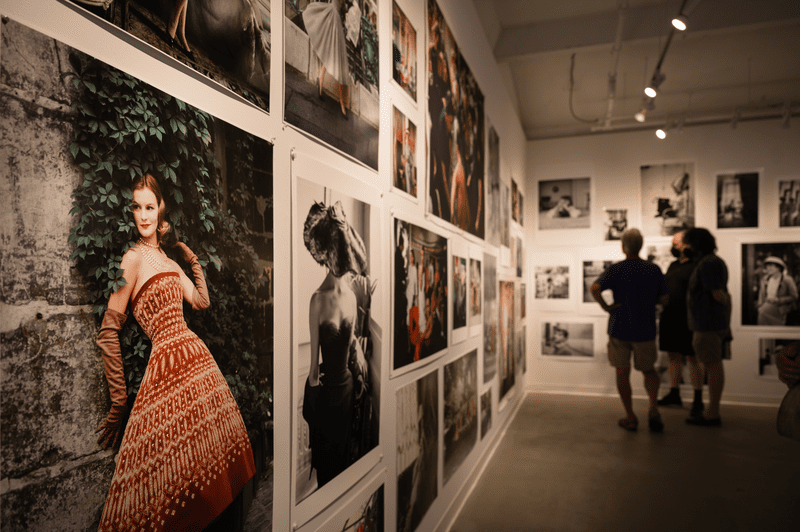 People in the far right of an image look at paintings on a wall in an indoor gallery.