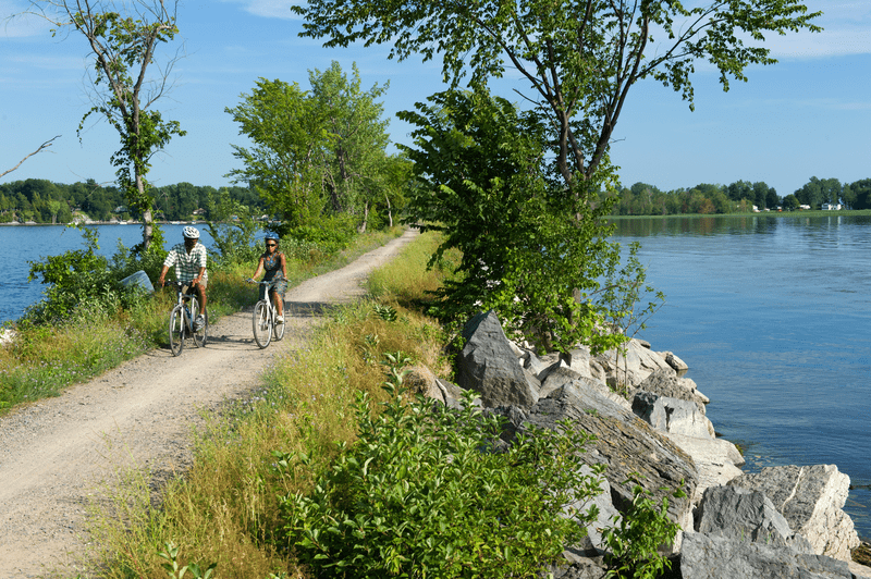 Two people ride bikes across a smooth gravel path that crosses a lake in the summer.