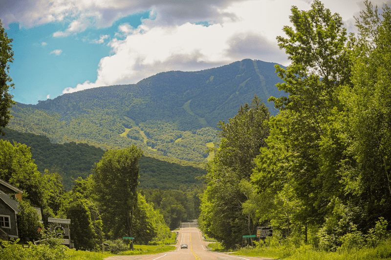 A car drives toward the camera on a road with green tree-covered mountains on both sides.