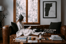 A person wearing a hat reads on a brown leather couch under a blanket. Outside, snowy trees are visible through a large window.