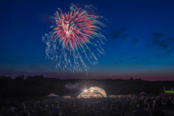 Fireworks streak across the sky above a lit stage at dusk.