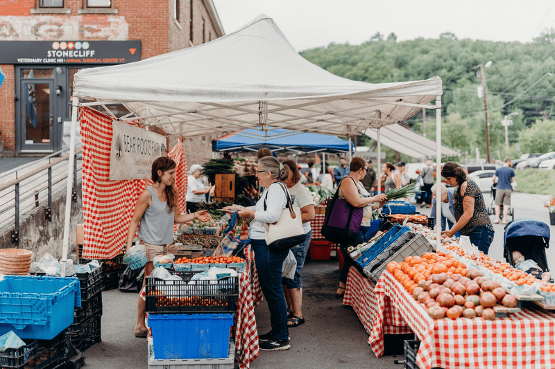 Groups of people shop produce and other goods under tents at an outdoor market.