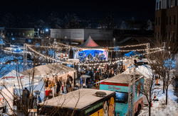 Food trucks and vendor tents line lit walking lanes, with a band is playing on a stage, outdoors in the winter.