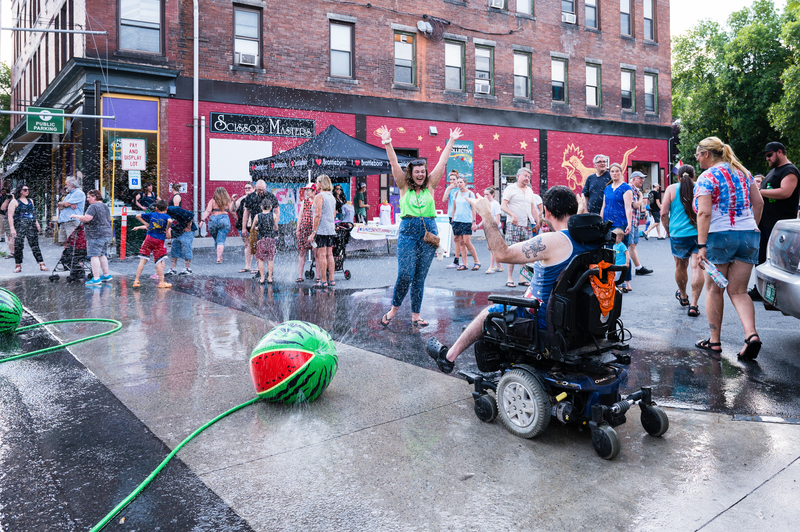 An outdoor celebration on a warm day with people in a paved parking lot, hoses spraying water, and vendors in the background.