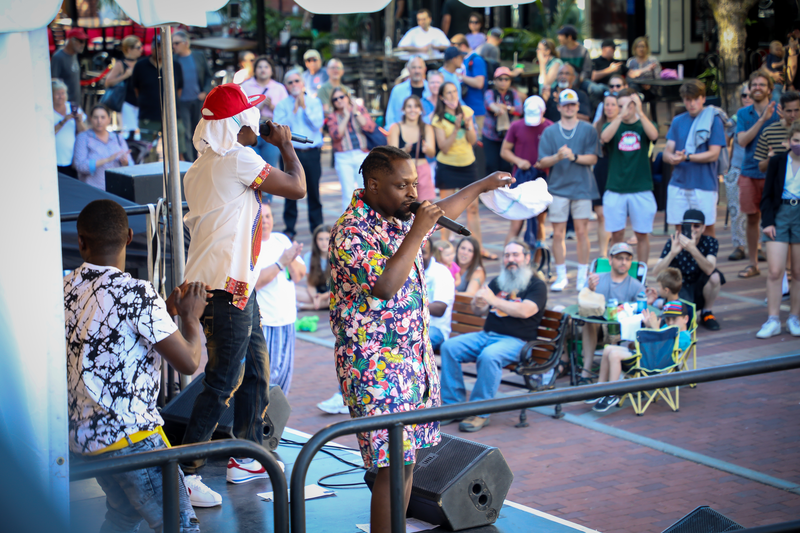 A crowd gathers around a person singing into a microphone in a street performance.