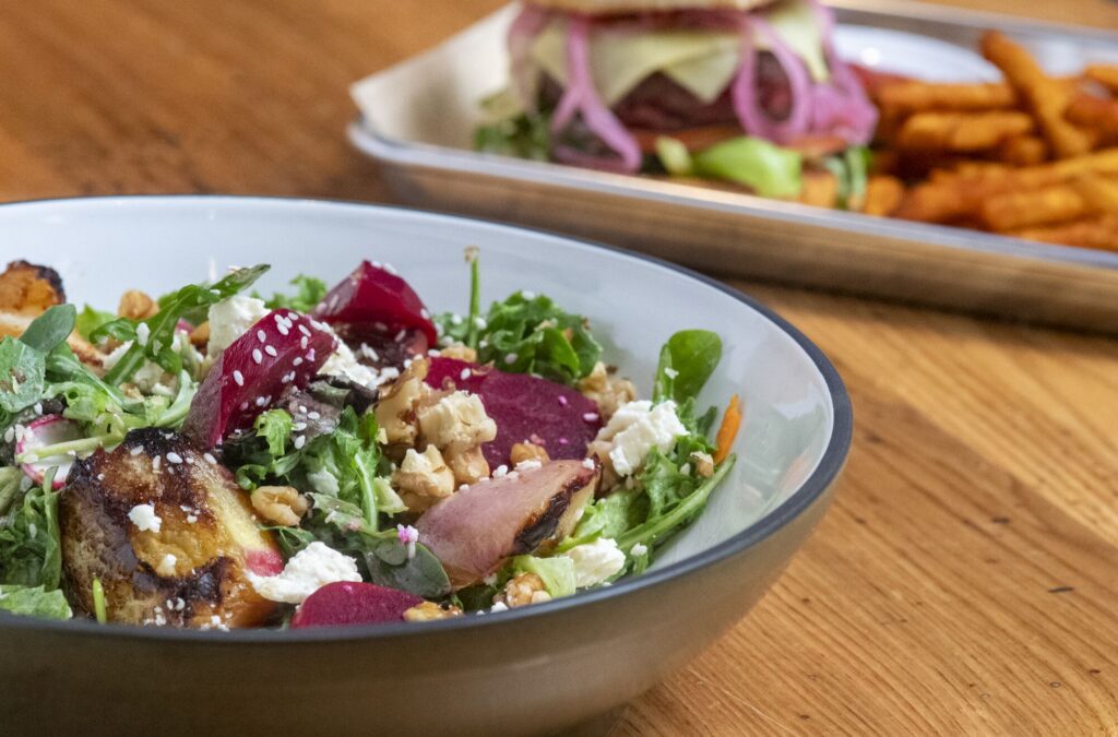 close up view of a colorful salad with a plate of burger and fries in the background.