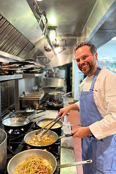 A chef poses for the camera while preparing a dish in a kitchen.