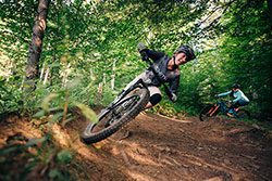 A couple is mountain biking around a banked, dirt path in the woods.