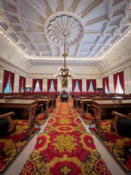 The inside of a legislative chamber, with red velvet drapes on the windows and ornate red carpets.