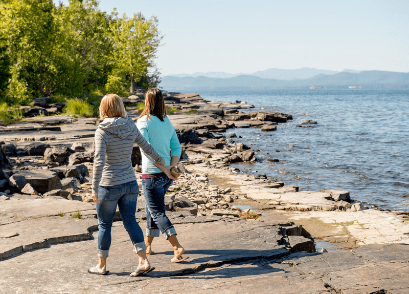 Two people walk and hold hands along next to a lake on a sunny day.