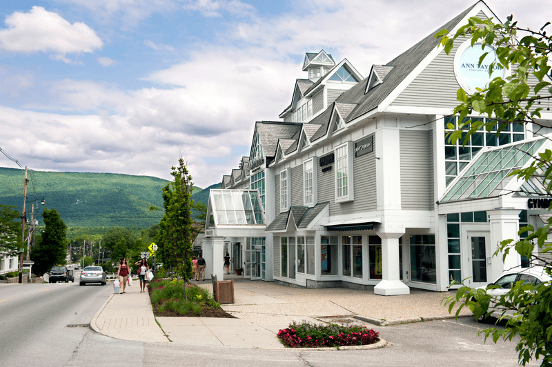 White buildings housing stores on a downtown street with green mountains behind. 