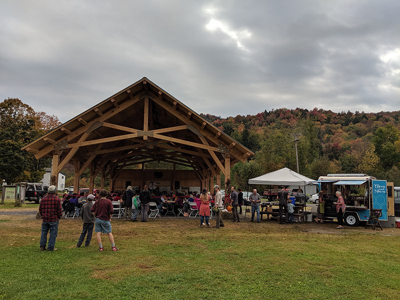 People gathered under a pavilion and in the grass with a tent and food truck.