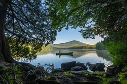 Seen from behind tree branches, a person paddles a canoe on a lake. A mountain is visible behind.