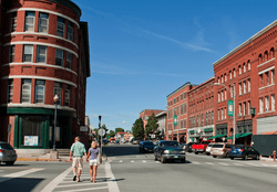 Two people walk down a street with large brown brick buildings on a warm sunny day.