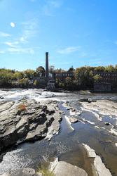 A wide and shallow river passes by an old smokestack that rises above the trees on the opposite bank.