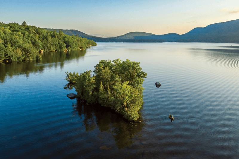 Ariel view of a large lake with a tree-covered island and surrounded by mountains.