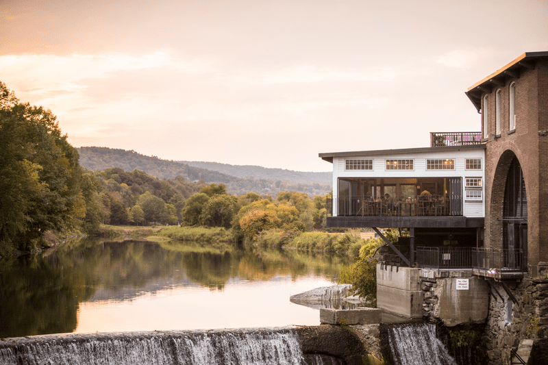 A historic building partially hangs over a calm flowing river with mountains in the background.