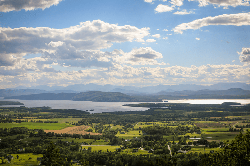 The view from the top of a mountain with fields, a lake, and more mountains visible.