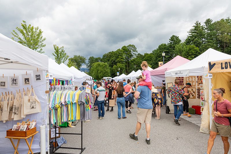 People walk between rows of tents with merchants selling their goods on a warm day.