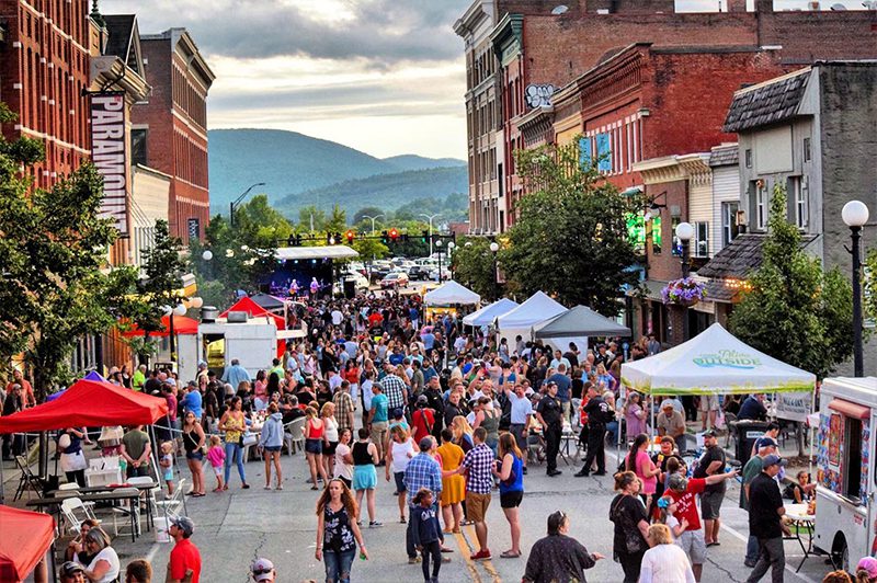 A crowd of people walk around a street lined tents. Mountains can be seen between brick buildings in the background.