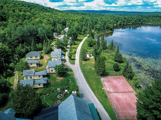 Seen from above, a resort set along a main road with a lake on the right side. It’s summer and the surrounding mountains are covered with green trees.