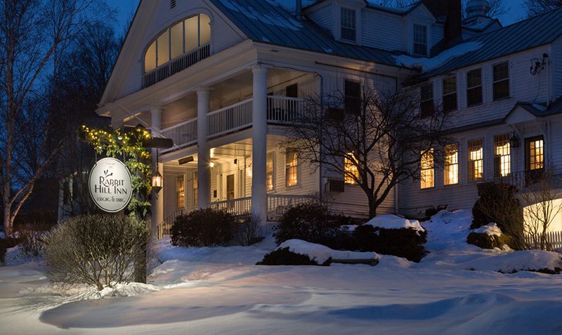 A hotel lit up at dusk with a sign reading Rabbit Hill Inn. Snow blankets the winter scene.