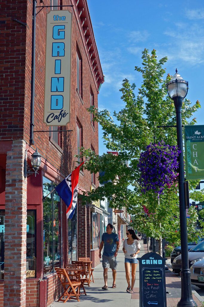 Two people walk along a sidewalk next to brick buildings with store fronts, and a sign that reads 