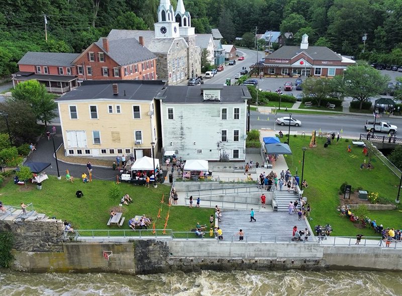 Seen from above, a small rural town adjacent to a flowing river and people in a green field.