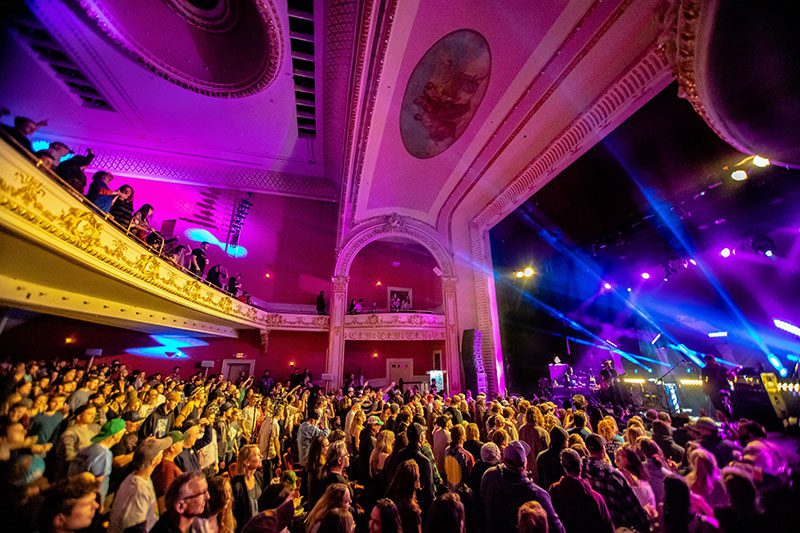 A crowd at an indoor concert with bright purple light streaming from the stage.