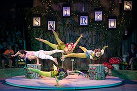 Four brightly dressed Circus performers on a stage, all balancing on one bent arm and facing in different directions.