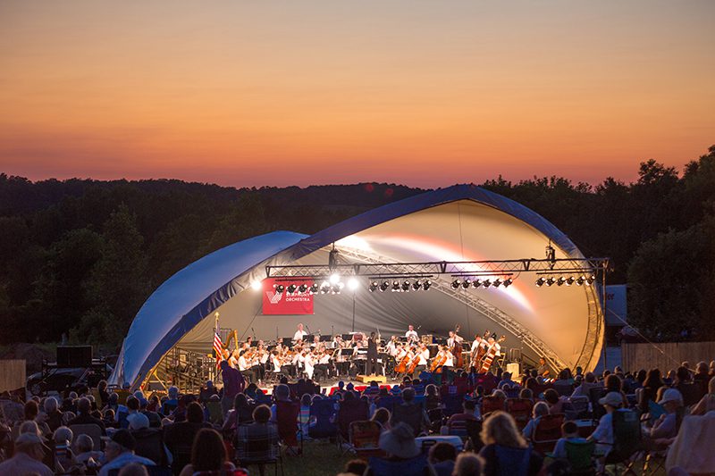 An audience sits down to watch an outdoor concert as the sun sets in the background.