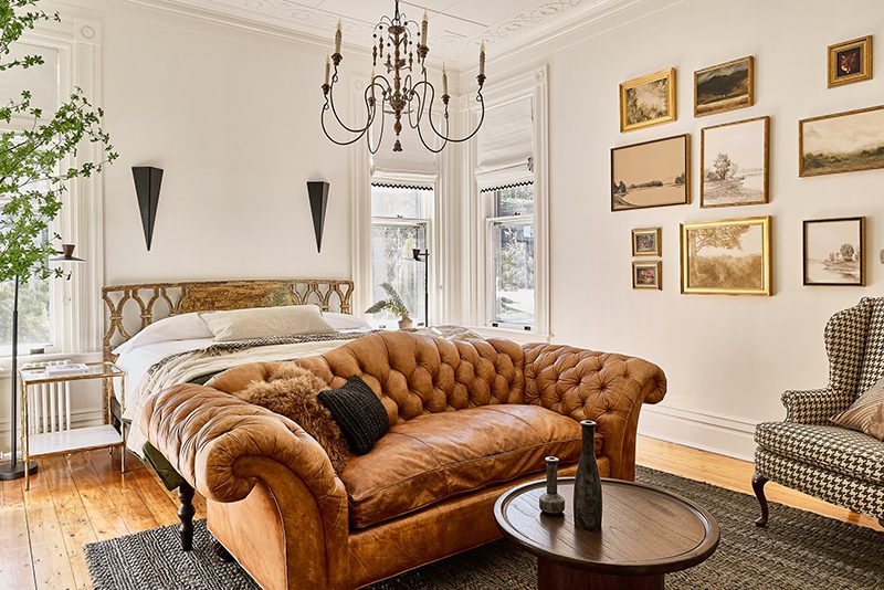 The interior of a luxurious hotel guest room with a chandelier hanging from the ceiling and a leather couch at the end of a bed.